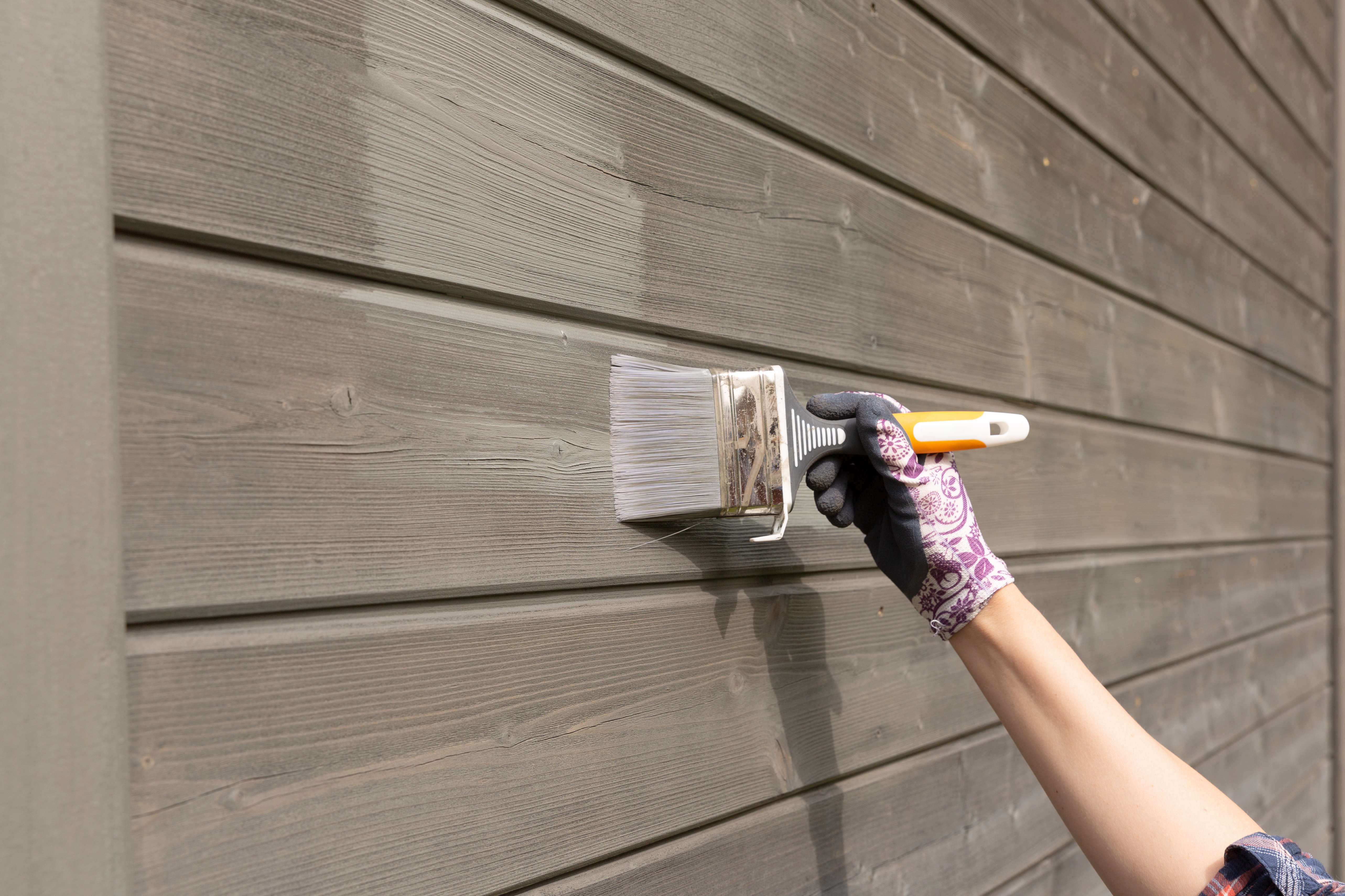 Worker painting wooden siding with brush and yellow-handled paintbrush