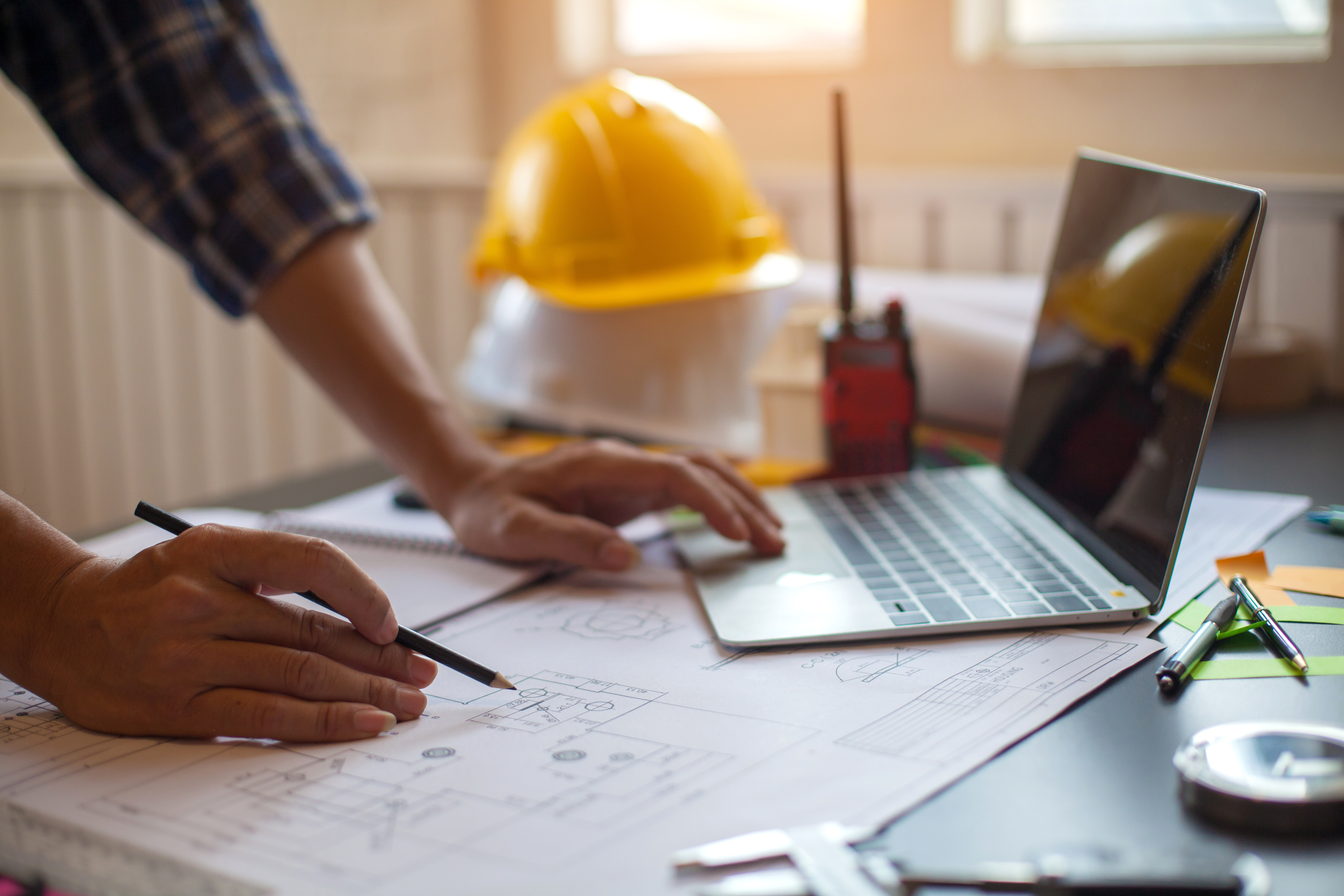 Construction worker reviewing blueprints at desk with hard hat and laptop
